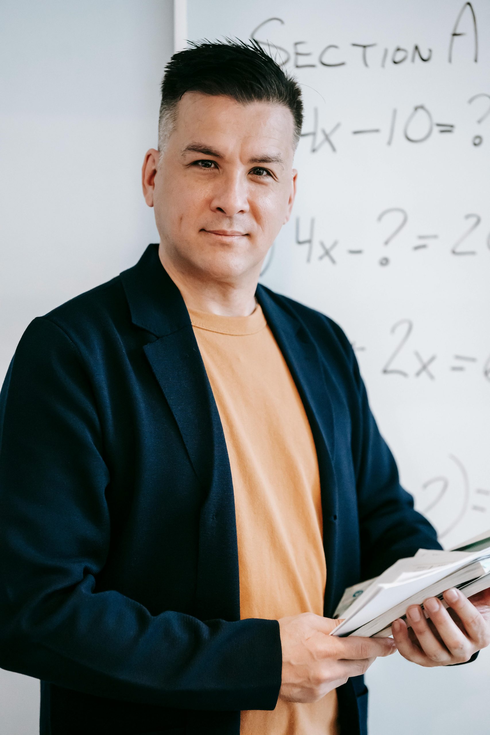 Male teacher standing in a classroom, holding books with a smile in front of a whiteboard.